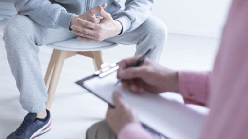 Kuvituskuva. Kuvan henkilöt näkyvät vain osittain. Toisella henkilöllä on kynä kädessä ja hän kirjoittaa paperille. Taustalla oleva henkilö istuu jakkaralla.