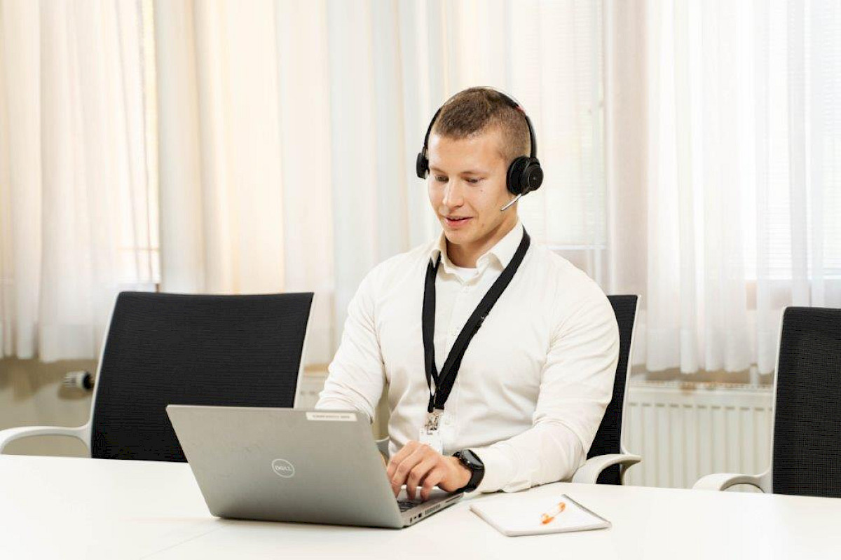 Photo of a male person wearing headphones and sitting at a desk working on a computer. Light window curtains in the background.
