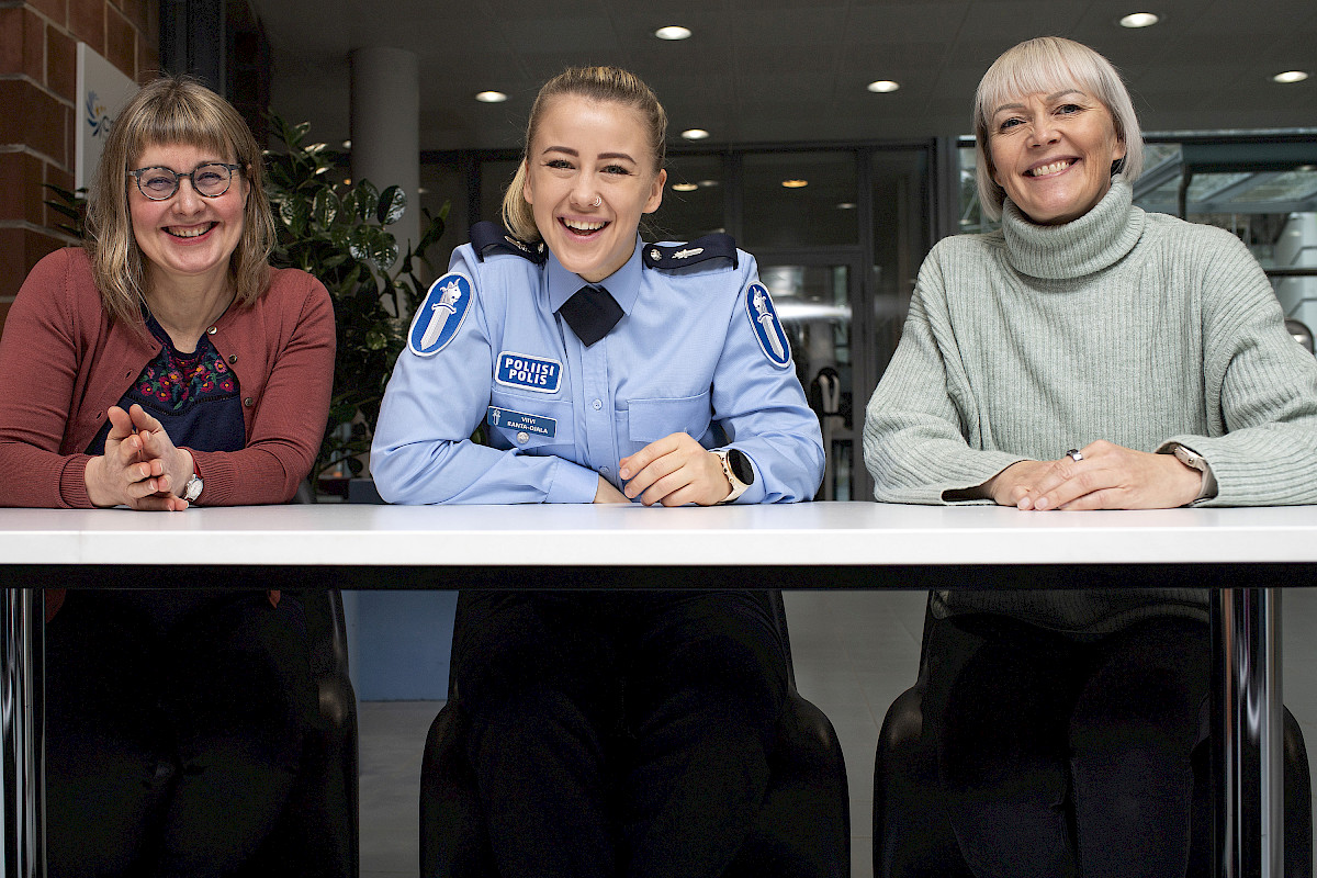 Kolme iloista naishenkilöä kuvattuna edestäpäin istumassa pöydän ääressä. Keskemmäisellä on poliisin uniformu ja kahdella muulla siviilivaatteet.