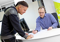 Illustration. There are two people at the table. One stands and the other sits behind the table. People look at the form on the table.