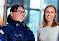 A group of people in a bright office environment. The group includes two police officers and two civilians. Police officers wear dark blue uniforms with the text 'POLICE' on the back. Civilians are dressed in casual clothes. People's faces are blurred for privacy. In the background is a large window and a table.