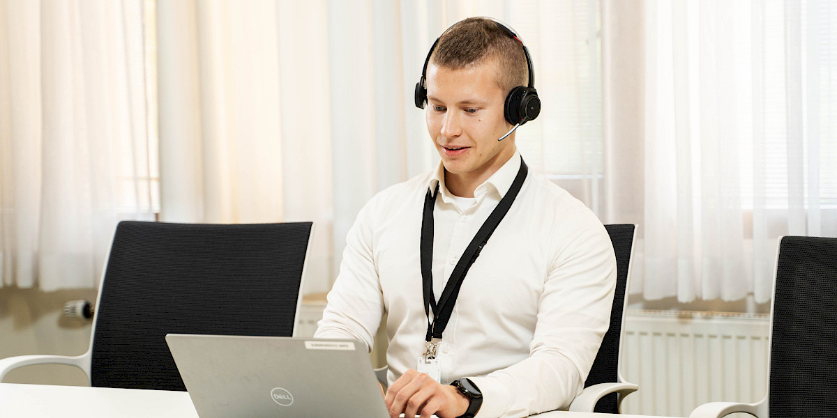 Photo of a male person wearing headphones and sitting at a desk working on a computer. Light window curtains in the background.