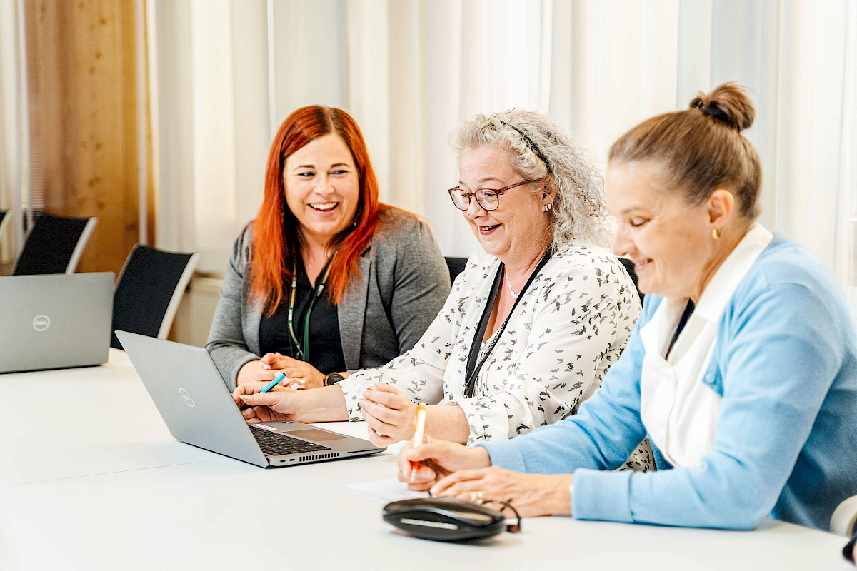 Three cheerful women sitting at a desk in a conference room. One has a computer in front of him. Light window curtains in the background.
